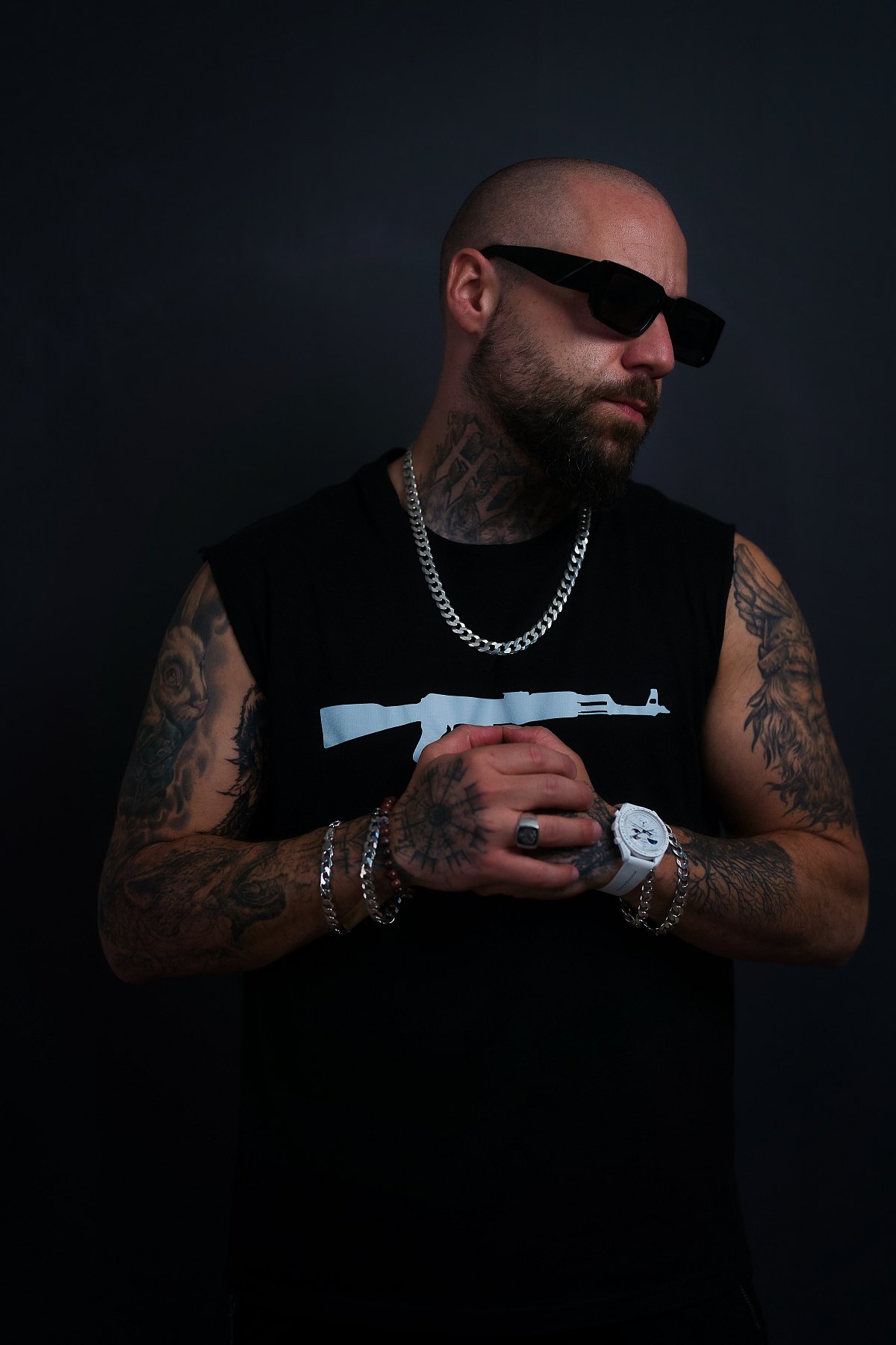 Tätowierter Mann mit Sonnenbrille, Bart und Silberkette steht vor dunklem Hintergrund und hält die Hände verschränkt.