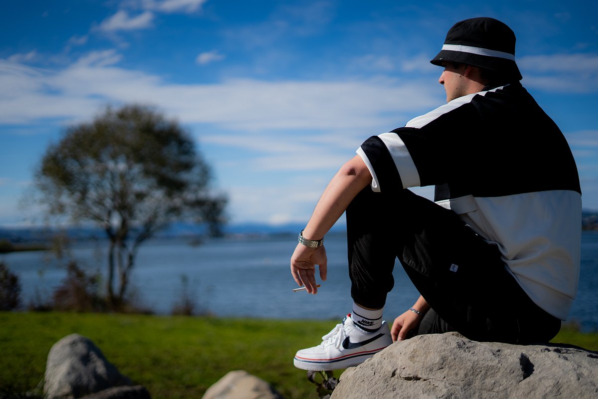 Person mit Bucket Hat sitzt auf einem Felsen am Seeufer und blickt bei blauem Himmel aufs Wasser.