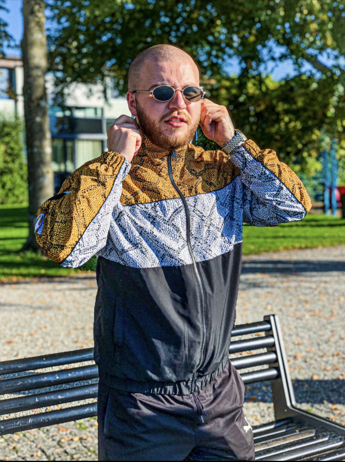 Mann in gemustertem Retro-Trainingsanzug mit schmaler Sonnenbrille posiert bei Sonnenschein vor einer Parkbank.