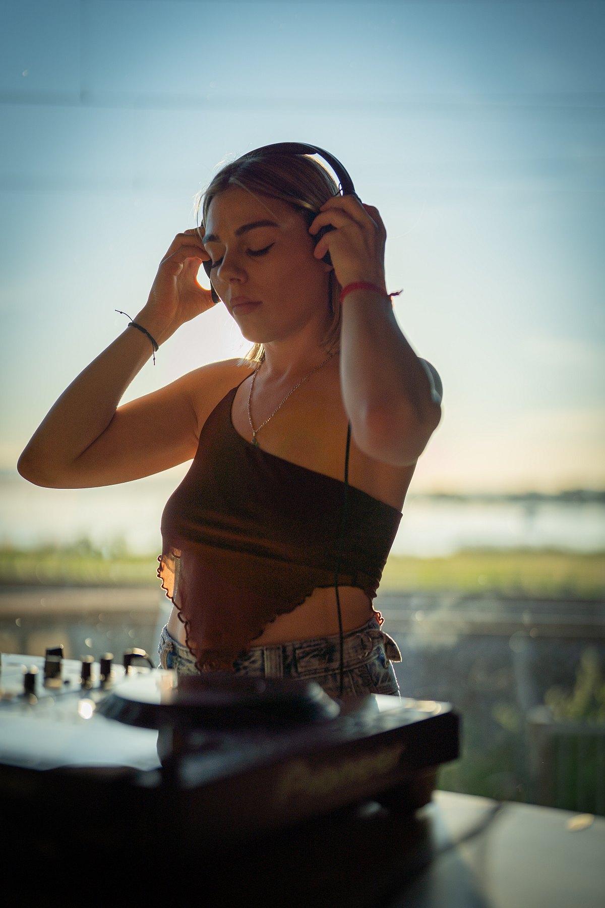 Junge Frau am DJ-Pult hält im Gegenlicht der Abendsonne die Kopfhörer an die Ohren und schliesst die Augen.