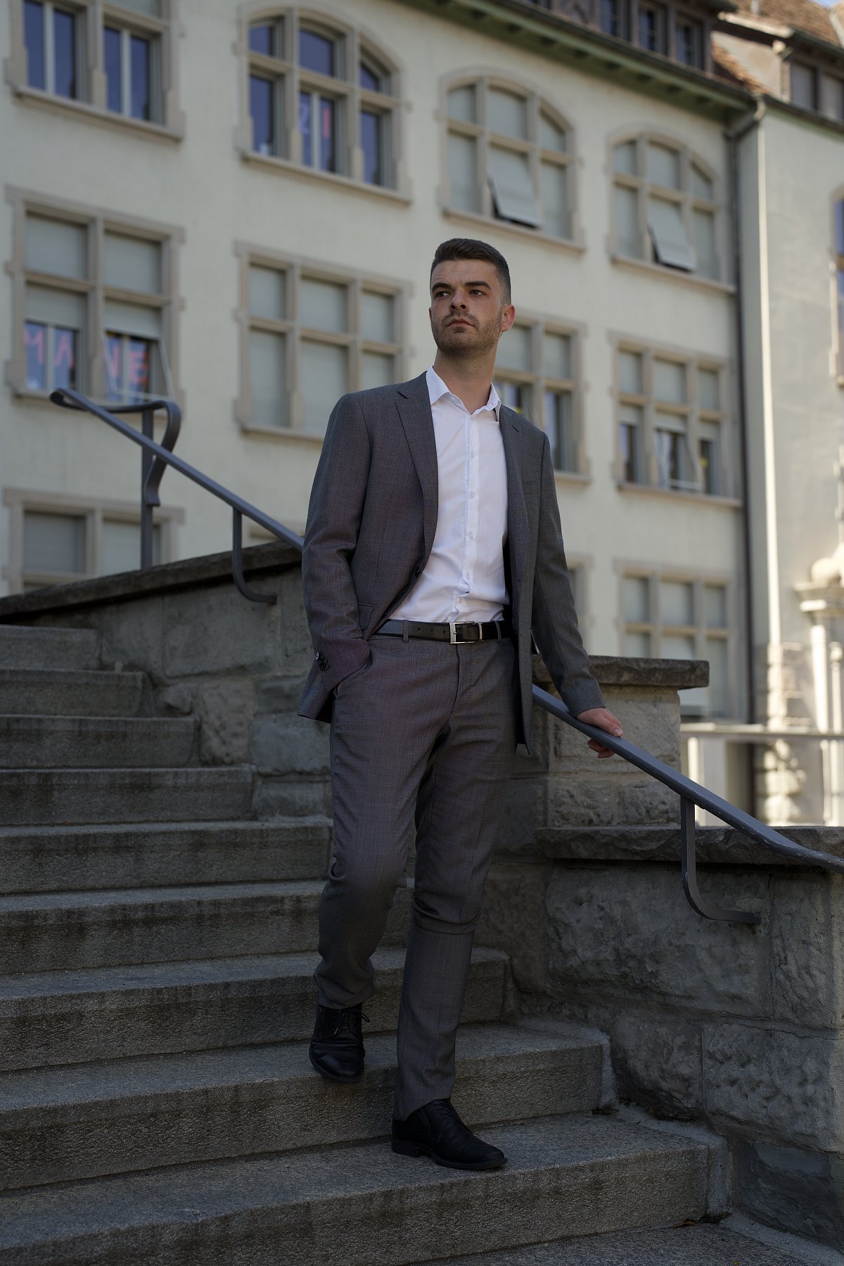 Junger Mann im grauen Anzug steht auf einer Steintreppe vor einer hellen Altbaufassade.