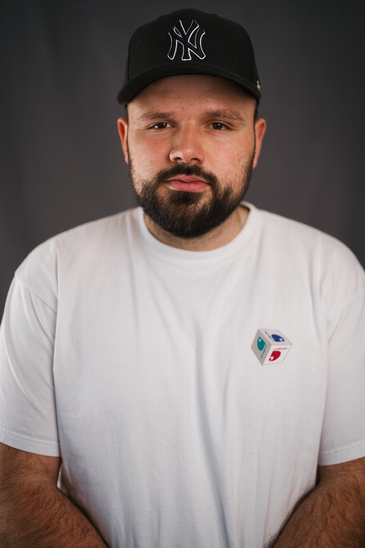 Junger Mann mit schwarzer Cap und Bart im weissen T-Shirt vor grauem Studiohintergrund.