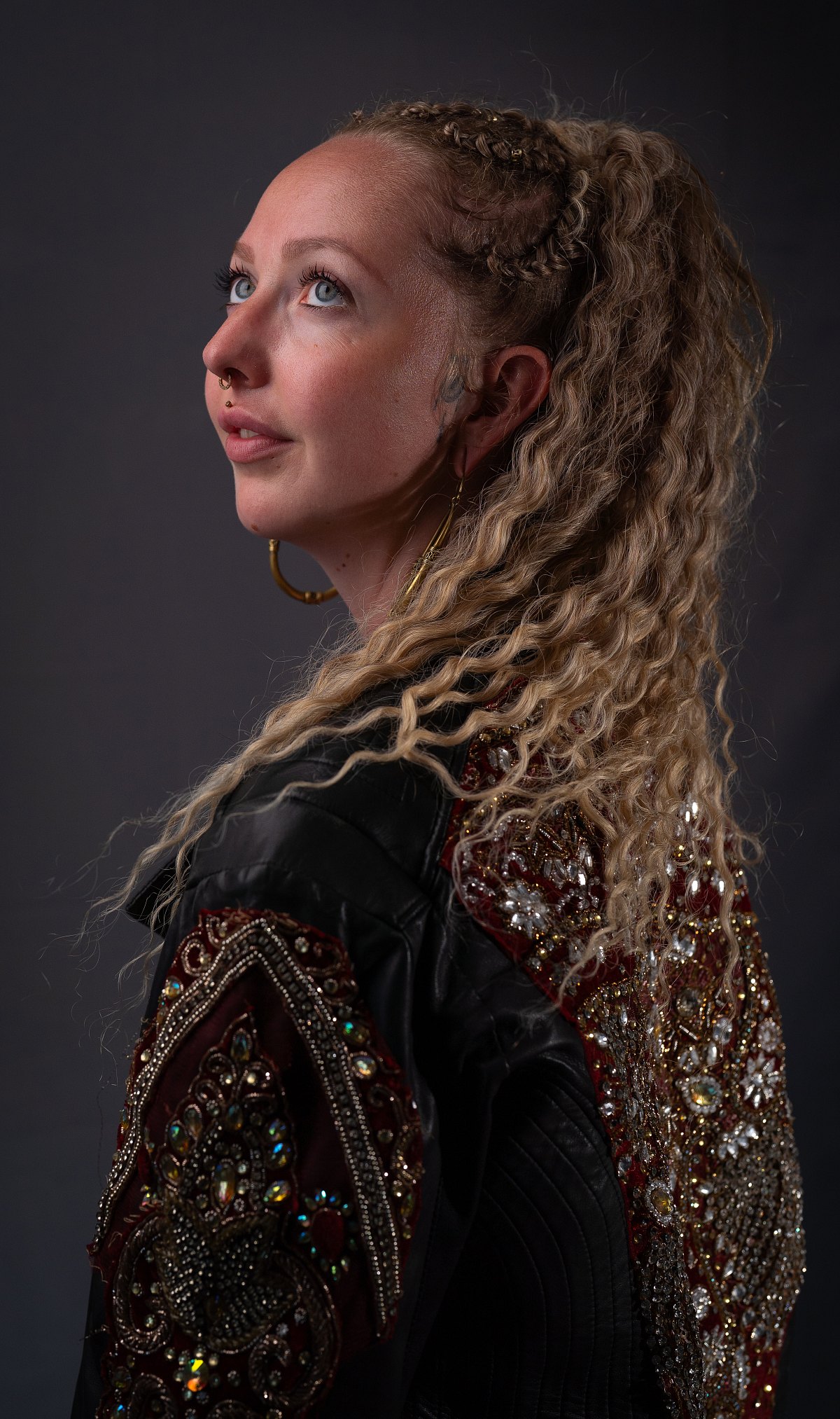 Studioporträt einer Frau mit langen blonden Locken und verzierter Lederjacke vor dunklem Hintergrund.