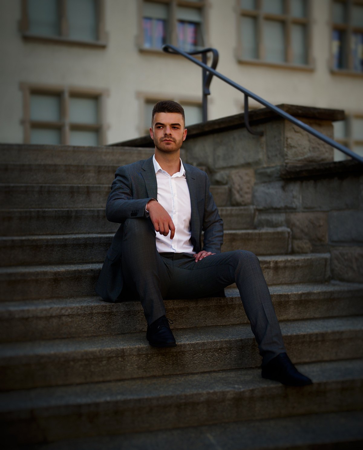 Junger Mann im grauen Anzug sitzt auf einer Steintreppe und blickt in die Kamera.