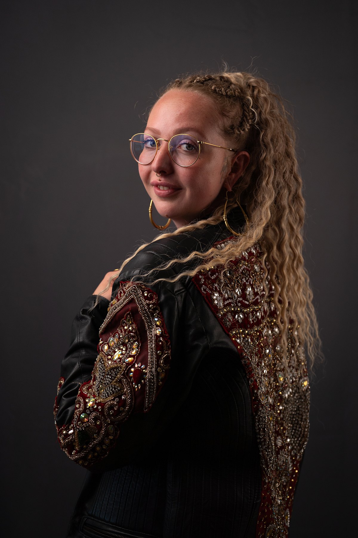 Junge Frau mit langen blonden Locken, runder Brille und bestickter Lederjacke blickt über die Schulter in die Kamera.
